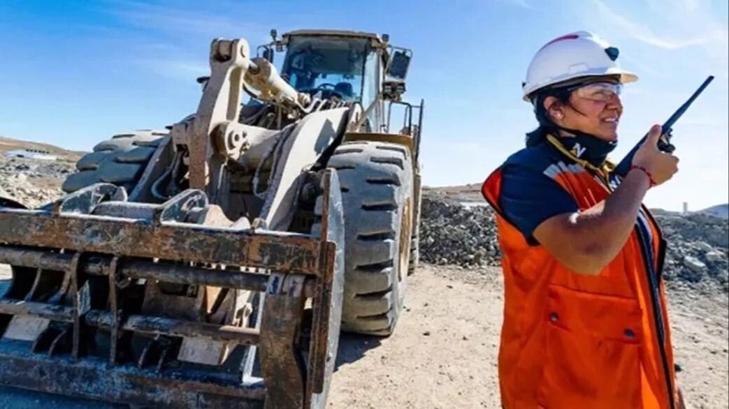 Trabajadora minera con casco y chaleco reflectante junto a una pala cargadora en un yacimiento