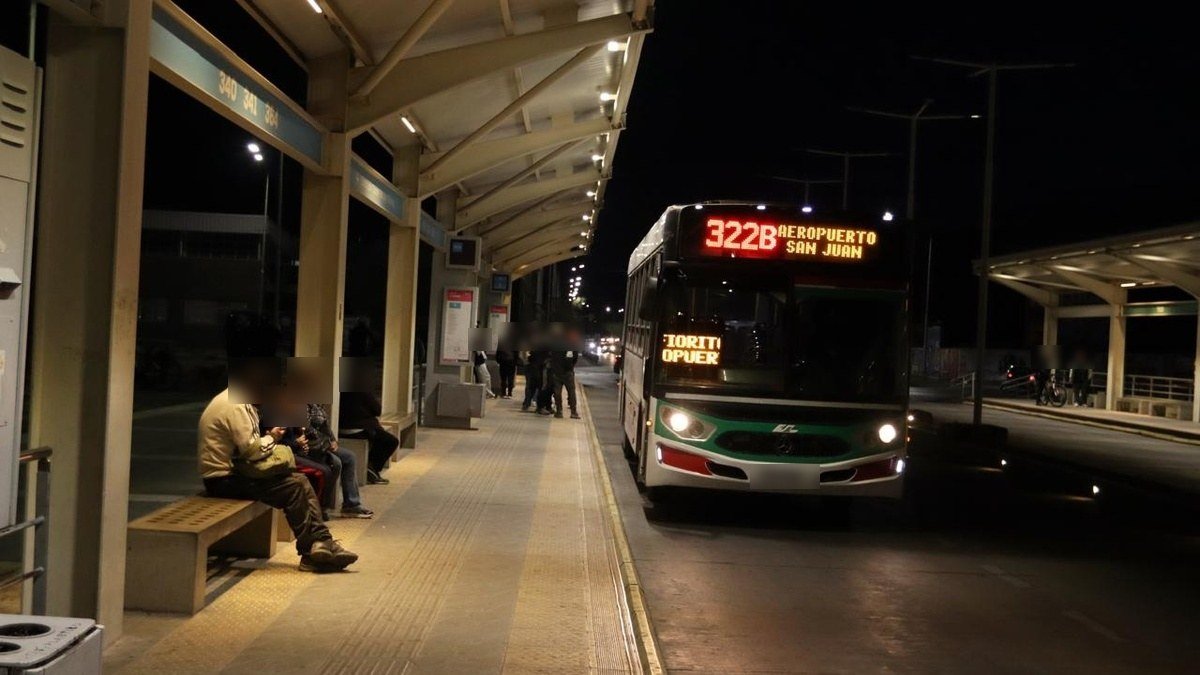 Colectivo de la línea 322B con destino Aeropuerto San Juan detenido de noche junto a una parada techada con pasajeros sentados