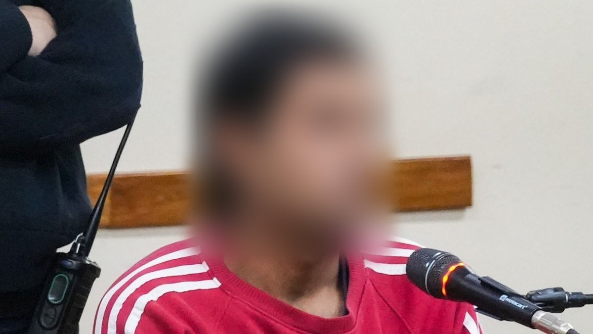 Hombre con buzo rojo sentado frente a un micrófono en una sala de audiencias, con el rostro difuminado