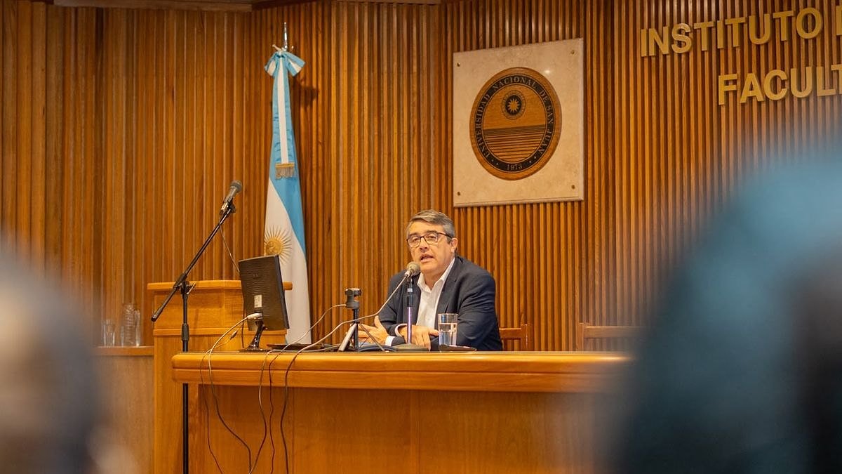 Marcelo Molina expone sentado en el estrado de un auditorio, junto a una bandera argentina