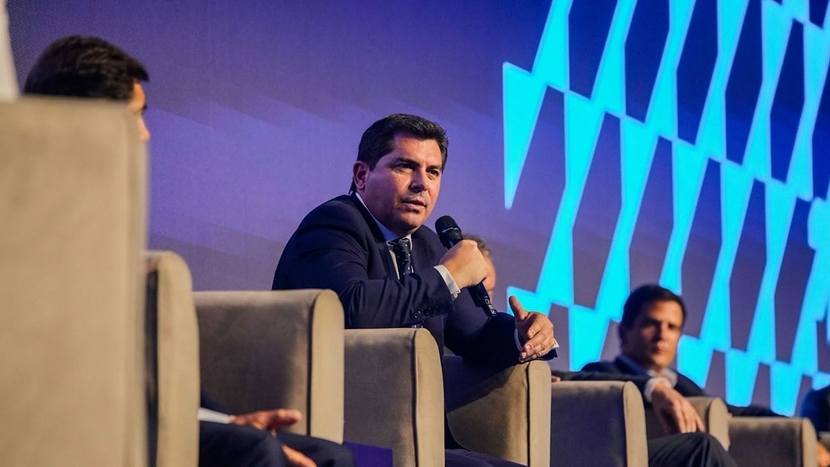 Marcelo Orrego habla con un micrófono, sentado en un panel frente a una pantalla azul