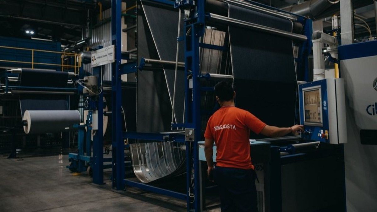 Un operario con remera naranja maneja el tablero de una máquina textil con rollos de tela