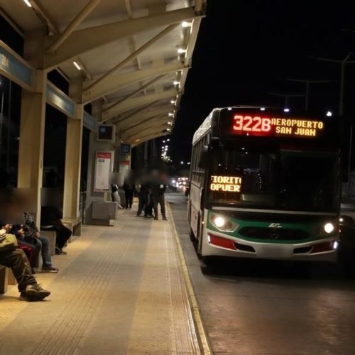 Colectivo de la línea 322B con destino Aeropuerto San Juan detenido de noche junto a una parada techada con pasajeros sentados