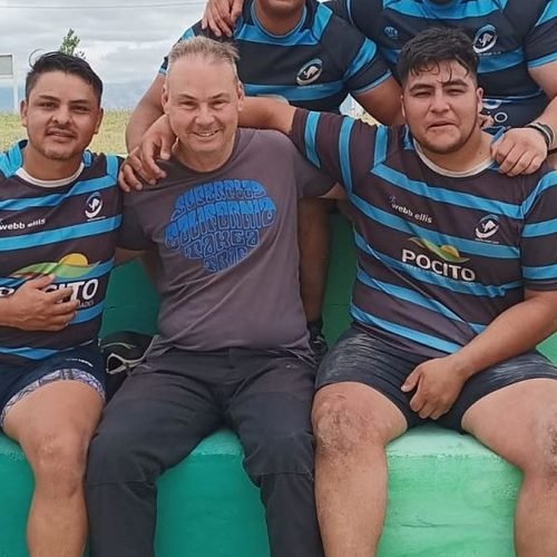 Cinco rugbiers con camisetas a rayas sentados en una tribuna de cemento verde junto a la cancha