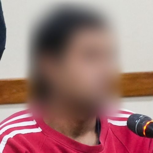 Hombre con buzo rojo sentado frente a un micrófono en una sala de audiencias, con el rostro difuminado