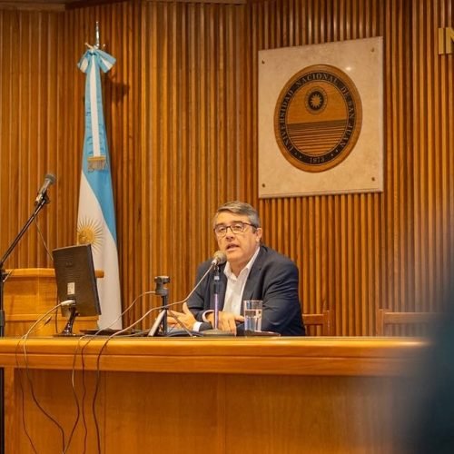 Marcelo Molina expone sentado en el estrado de un auditorio, junto a una bandera argentina