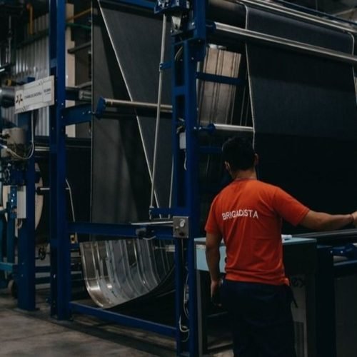 Un operario con remera naranja maneja el tablero de una máquina textil con rollos de tela